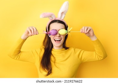 Funny Young Caucasian Woman Covers Eyes With Two Easter Eggs, Wears Fluffy Bunny Ears, Dressed In Sweater, Model On Yellow Background With Free Space For Promotion. Spring Holiday And Easter Concept