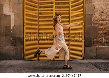 Funny young caucasian woman looks at camera, runs along street in warm weather on weekend. Blonde wears beige sundress and black sandals. Summer vacation concept