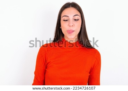 Funny Young caucasian woman wearing red sweater over white background makes grimace and crosses eyes plays fool has fun alone sticks out tongue.