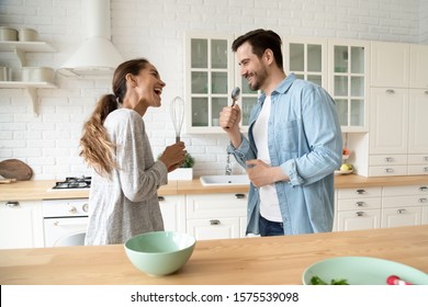 Funny Young Couple Singing In Kitchenware Microphones In Kitchen, Happy Carefree Husband And Wife Hold Beater Spoon Together Having Fun Preparing Dinner Breakfast Cooking Dancing Listen Music At Home