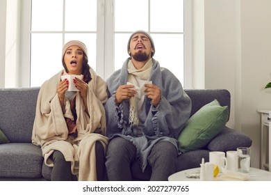 Funny young couple in warm hats, wrapped in blankets, sitting together on couch at home and sneezing in paper tissues. Sick husband and wife suffering from unpleasant symptoms of bad cold or flu virus