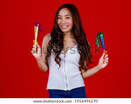 Funny young and cure Asian girl holding and ready to blow playing party popers with funny and happy. Studio shot on red background.