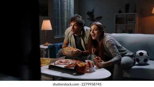 Funny young emotional caucasian sport fan couple watching a game together, eating popcorn and pizza and reacting to their team playing  - Powered by Shutterstock