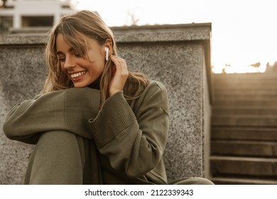 Funny young fair-skinned girl smiles broadly in headphones outdoors. Happy woman sitting on concrete staircase and listening to music. Relaxation and health care concept.