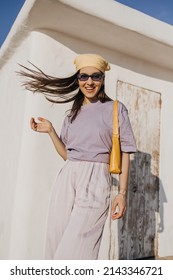 Funny young fair-skinned woman playing with her hair tied with handkerchief in open area. Brown-haired model in sunglasses wears T-shirt, pants and bag. Summer time concept