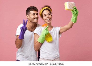 Funny young family fooling around while doing housework together: cheerful man and woman in rubber gloves posing for selfie, pretending that sponge is smart phone and smiling broadly at camera
