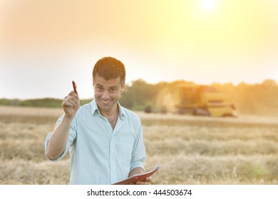 Funny young farmer with raised arm standing on wheat field with combine harvester in background