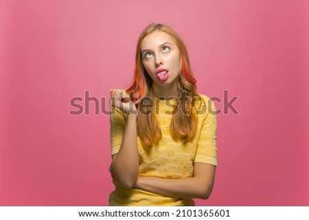 Funny young girl grimacing crossing eyes showing tongue, making foolish face expression, fooling on pink background