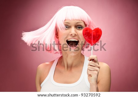 Funny young girl in pink wig holding lollipop and posing for camera across pink background