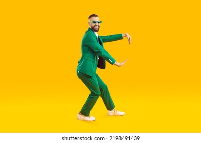 Funny Young Guy Dancing In A Cool Party Outfit. Happy, Joyful Man Wearing A Stylish Green Suit And Sunglasses Dancing Isolated On A Bright Yellow Colour Background. Full Body Studio Shot, Side View