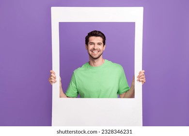 Funny young guy photographing through big white frame cadre memories capture wearing trendy t-shirt isolated on violet color background