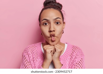 Funny young Latin woman with dark hair puckers lips keeps hands under chin makes fish face funny grimace wears casual clothing poses against pink background. Human facial expression concept.