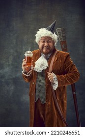 Funny Young Man In Brown Vintage Suit And White Wig Like Medieval Royal Hunter With Old Gun Laughing Isolated On Dark Background. Comparison Of Eras Concept, Renaissance Style.