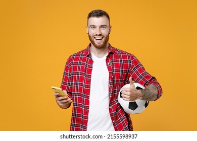 Funny young man football fan in red shirt cheer up support favorite team with soccer ball using mobile cell phone showing thumb up isolated on yellow background studio. People sport leisure concept