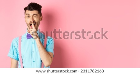 Funny young man planning a surprise, hushing and winking at camera, asking to keep quiet, telling a secret, standing on pink background.