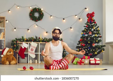 Funny young man in Santa cap and holiday costume sitting on fitness carpet and meditating over decorated home with Christmas tree at background. Christmas and New Year at home concept