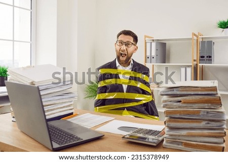 Funny young man, stuck at work in office and bound with yellow scotch tape, sitting at his desk with laptop computer and piles of paper folders, screaming loudly and asking for freedom