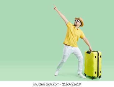 Funny young man in summer clothes going on holiday trip and having fun. Happy excited tourist with yellow suitcase showing something on blank empty green copy space background