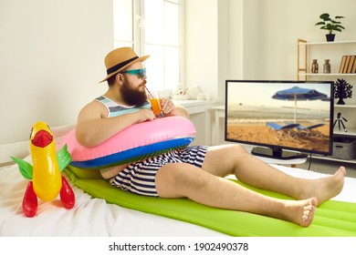 Funny young man with sunglasses and inflatable beach toys sipping cocktail and watching travel show on TV. Concept of canceled summer holiday plans, vacation in lockdown at home or Covid-19 quarantine - Shutterstock ID 1902490378