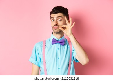 Funny Young Man Touching His French Moustache And Pucker Lips, Looking Silly At Camera, Standing In Bow-tie And Suspenders On Pink Background