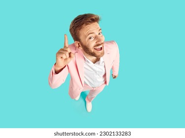 Funny young man in trendy pink suit standing isolated on blue background has good, clever, genius idea, looks at camera with happy face expression, and points his forefinger up. High angle view shot