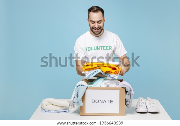 Funny young man in white volunteer t-shirt\
standing near table packing clothes in donation box for needy\
isolated on blue background studio. Voluntary free work assistance\
help charity grace\
concept