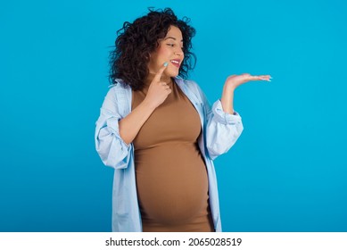 Funny Young Pregnant Arab Woman Standing On Blue Studio Background Holding Open Palm New Product. I Wanna Buy It!