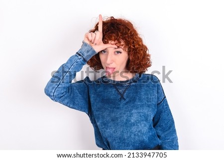 Funny young redhead girl wearing blue sweater over white background makes loser gesture mocking at someone sticks out tongue making grimace face.