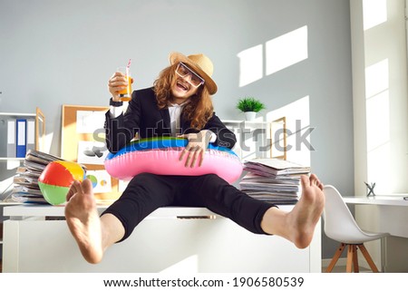Funny young smiling man accountant financial worker with long hair in hat sitting on office table with cocktail and dreaming of tropical vacations and beach. Crazy funny accountant worker concept