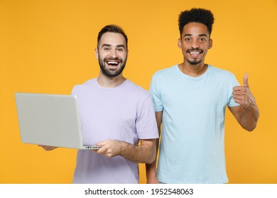 Funny young two friends european african american men 20s in violet blue casual t-shirts working on laptop pc computer showing thumb up isolated on bright yellow colour background studio portrait