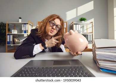 Funny Young Weird Man Accountant Financial Worker In Glasses With Long Hair Pointing At Piggy Bank With Finger In Office Near Laptop. Crazy Funny Accountant Concept