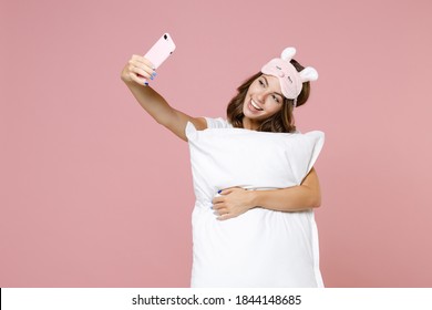 Funny young woman 20s in pajamas home wear sleep mask hold pillow doing selfie shot on mobile phone while resting at home isolated on pastel pink background studio portrait. Relax good mood concept