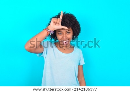 Funny Young woman with afro hairstyle wearing blue T-shirt over blue background makes loser gesture mocking at someone sticks out tongue making grimace face.