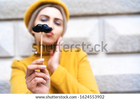 Funny young woman in a bright yellow trendy coat autumn winter 2020. Stands with a black lollipop in the shape of a male mustache and eyes wide open. Close-up portrait