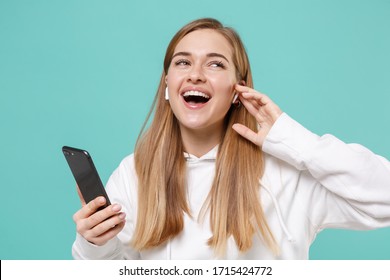 Funny Young Woman In Casual White Hoodie Posing Isolated On Blue Turquoise Wall Background Studio Portrait. People Lifestyle Concept. Mock Up Copy Space. Listen Music With Air Pods Using Mobile Phone