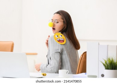 Funny young woman with clown nose and paper emoticon attached to her shoulder in office. April fools day celebration