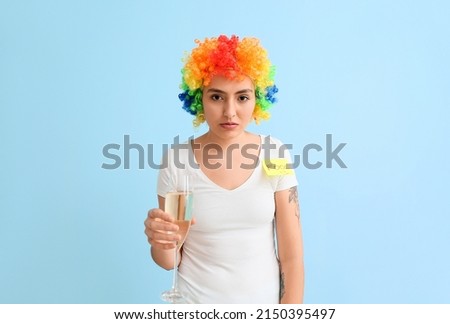 Funny young woman in colorful wig and with champagne on blue background. April fools' day celebration