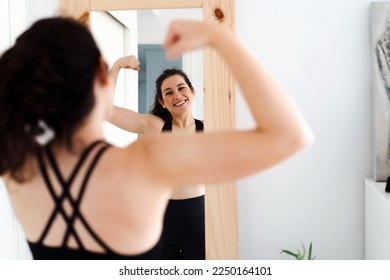 Funny young woman flexing arms looking in the mirror after doing her training - Shutterstock ID 2250164101