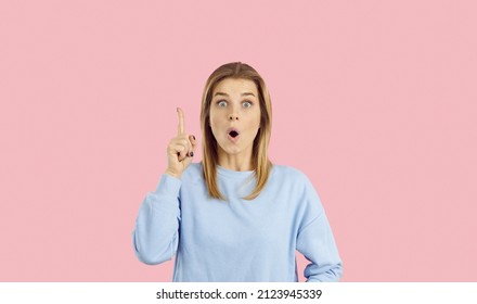 Funny Young Woman Has A Brilliant Idea. Happy Excited Beautiful Girl Isolated On Pastel Pink Having An Aha Moment, Pointing Her Index Finger Up And Looking At You With A Surprised Face Expression