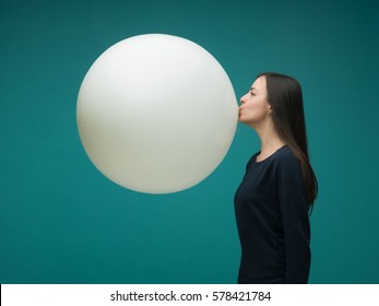 funny young woman with long dark hair inflating huge balloon on blue background