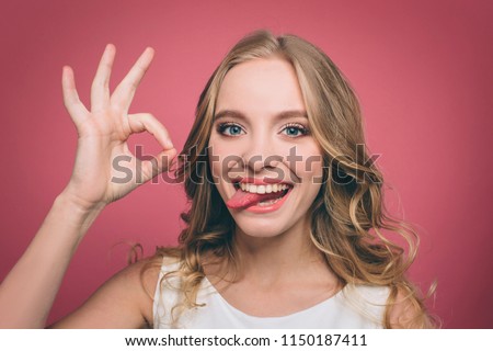 Funny young woman is looking straight forward and showing a piece of her tounge while she is smiling. Also girl is showing the ok symbol with her fingers. Isolated on pink background