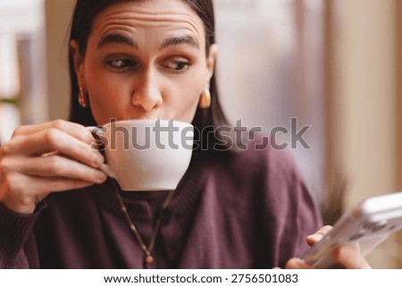 Funny young woman multitasking while drinking a cup of hot coffee and looking at her smartphone with a curious facial expression during a busy morning break in a cozy cafe interior