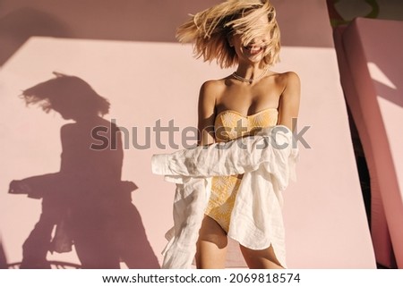 Funny young woman stands in yellow two-piece swimsuit waving her hair. Her shadow falls on pink wall from behind, blonde in shirt with smile on face. Happiness summer lifestyle concept.