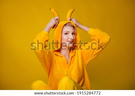 A funny young woman in yellow pajamas sits on a yellow background and holds bananas in her hands. Cheerful little girl fooling around with fruits. Studio photo