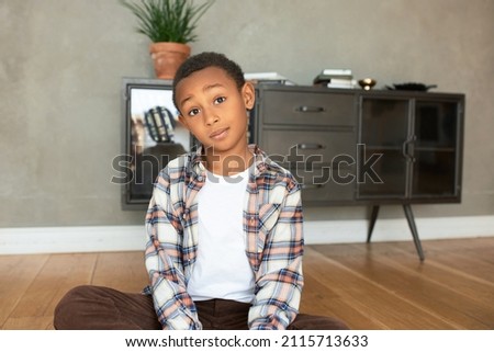 Funny-looking dark-skinned boy sitting on floor in living-room dressed in trendy flannel shirt and brown velvet jeans looking at camera with surprised face expression, raising eyebrows