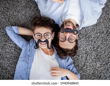 Funtime together. Curly father and son playing with fake moustache on sticks, lying on floor, top view - Powered by Shutterstock