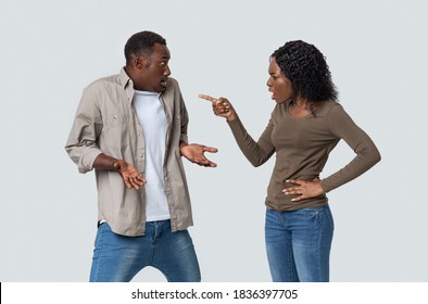 Furious African American Young Woman Yelling And Pointing At Her Innocent Boyfriend. Angry Black Lady Suspecting Her Husband In Cheating, Grey Studio Background. Distrust In Relationships