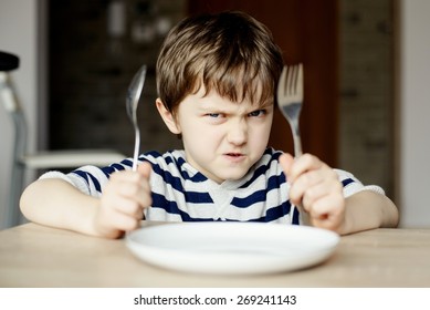 Furious Little Boy Waiting For Dinner. Holding A Spoon And Fork In The Hand