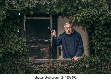 Furious Senior Man Gesturing With A Gun Protecting His Property, Standing In Front Of Old Overgrown Cabin.