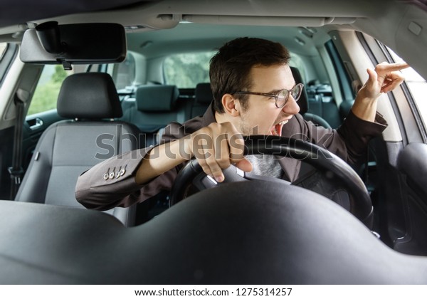 Furious young driver quarrels with other\
drivers. His right hand is on the steering\
wheel.